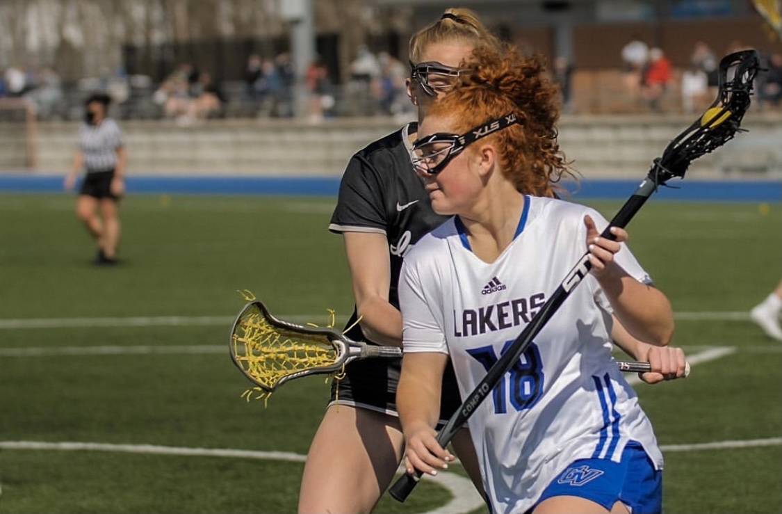 Active shot of a women's lacrosse player, number 18 wearing a white and blue uniform making a move on a defender